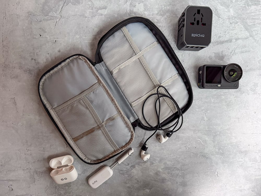 A selection of Ashley Wali's favorite travel tech in an FYY travel tech organizer. It includes Airpods and a CobCobb Ultra Strong Magnetic AirPod Pro Strap, an Twelve South AirFly Pro, an EPICKA Universal Travel Adapter, and a DJI Osmo Action 6 camera.