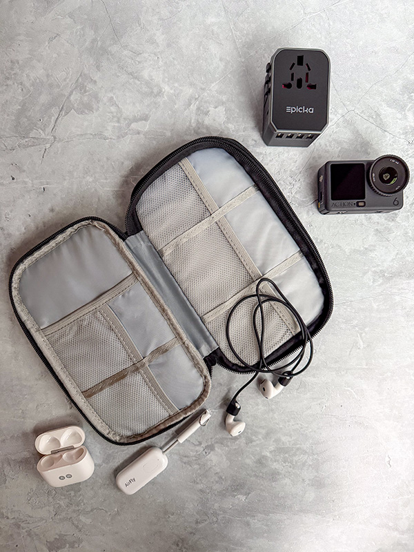 A selection of Ashley Wali's favorite travel tech in an FYY travel tech organizer. It includes Airpods and a CobCobb Ultra Strong Magnetic AirPod Pro Strap, an Twelve South AirFly Pro, an EPICKA Universal Travel Adapter, and a DJI Osmo Action 6 camera.